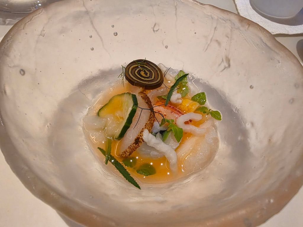 A beautifully arranged seafood dish in a textured, clear bowl featuring various types of raw fish, garnished with herbs and a decorative element.