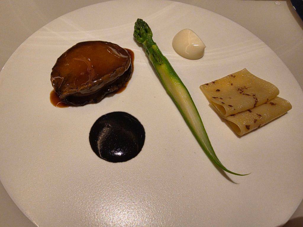 A gourmet plate featuring a piece of braised meat, accompanied by a green vegetable, a dollop of sauce, and roll of pasta on a white plate.