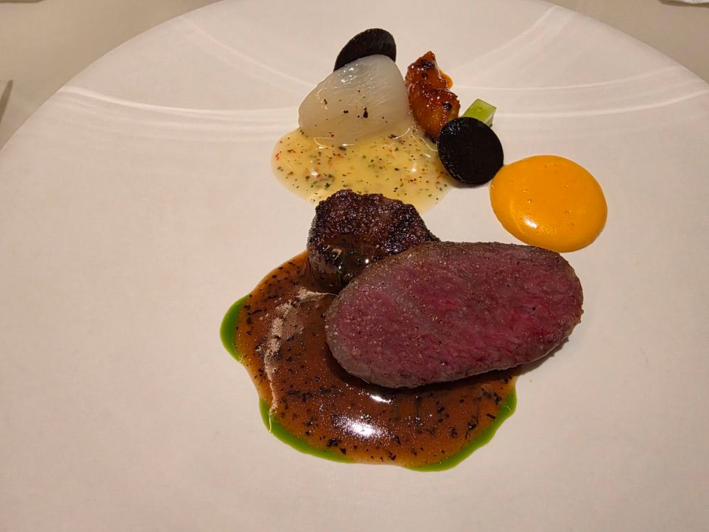 A gourmet dish featuring a perfectly cooked piece of beef, accompanied by various garnishes including a yellow sauce, grilled vegetables, and a green herb sauce, served on a white plate.