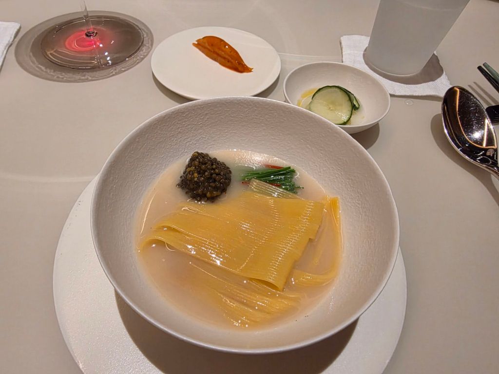 A gourmet dish featuring thin yellow noodle-like strands in a clear broth, topped with caviar and green herbs, accompanied by a side of pickled cucumber and a small serving of sauce.
