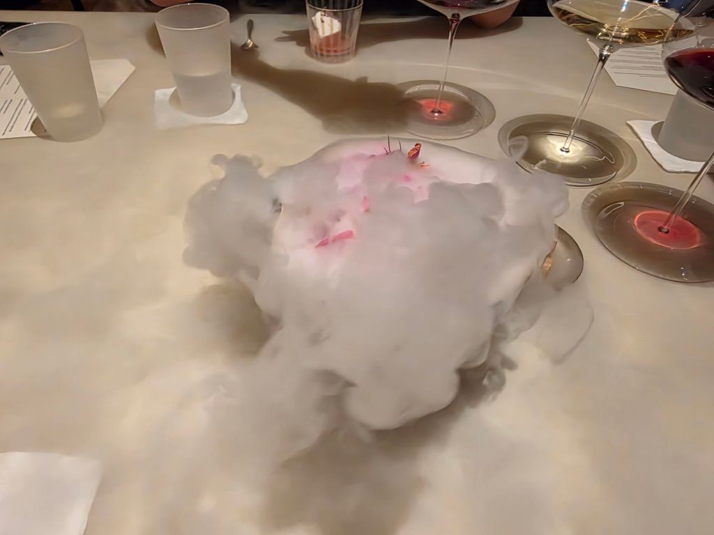 A dish surrounded by swirling mist, with decorative elements on top, and wine glasses visible in the background.