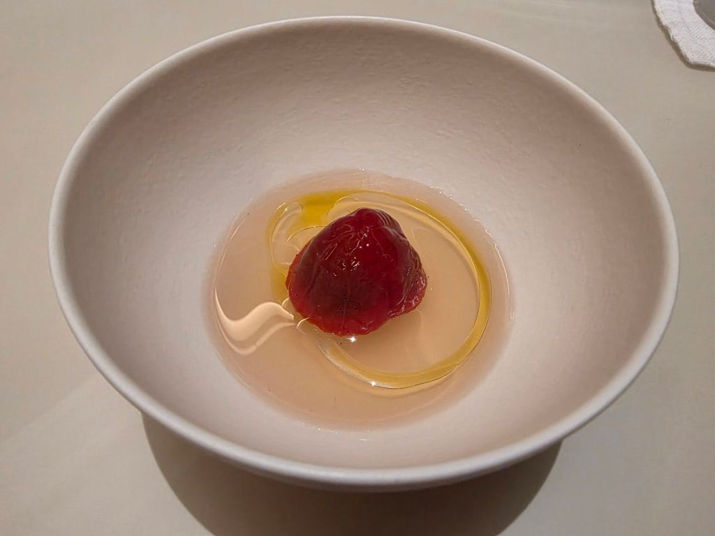 A white bowl containing a gelatinous dessert with a red center, surrounded by a clear liquid and a drizzle of yellow sauce.