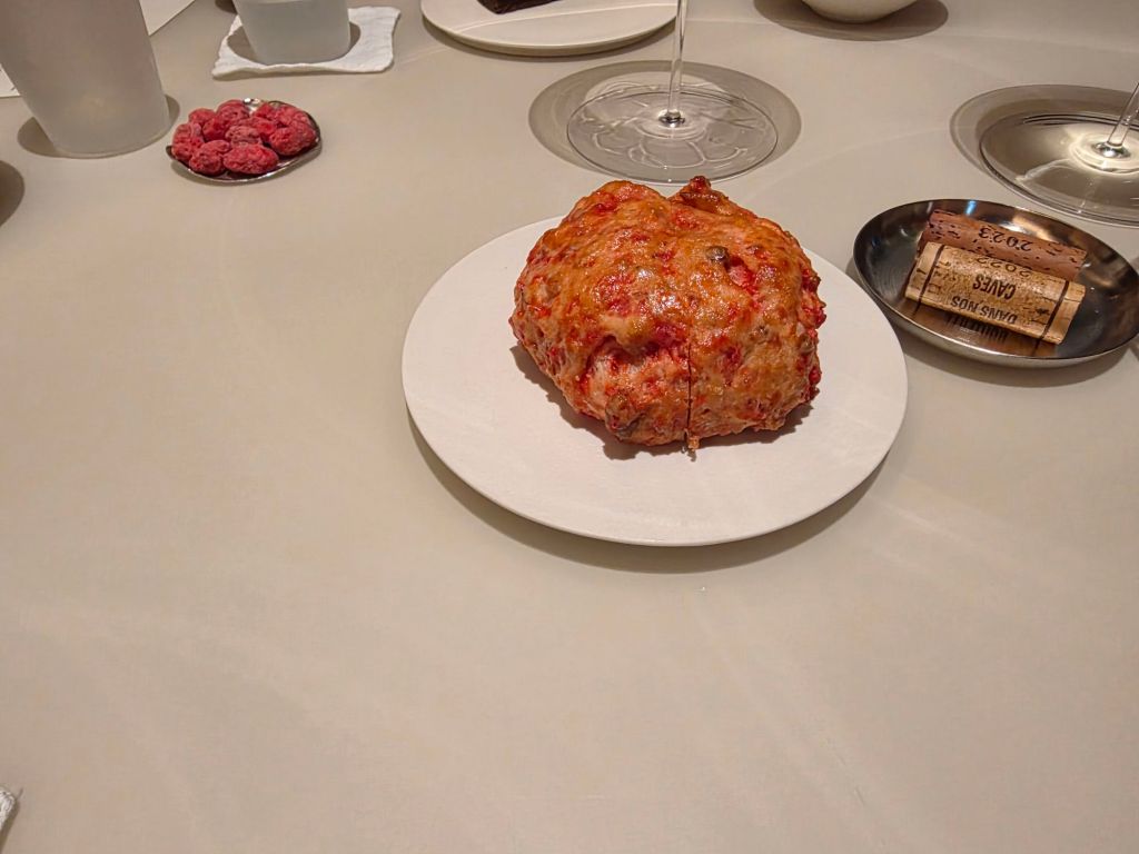 A close-up view of a round, baked dish placed on a white plate, with a golden-brown crust and visible ingredients resembling a savory meatloaf, surrounded by various dining items on a simple table setting.