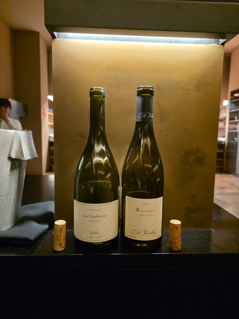Two wine bottles displayed on a black counter with a silver wine bucket in the background. The left bottle is labeled 'La Violette 2022' and the right bottle is labeled 'Bourgogne 2023'. Two corks are placed in front of the bottles.