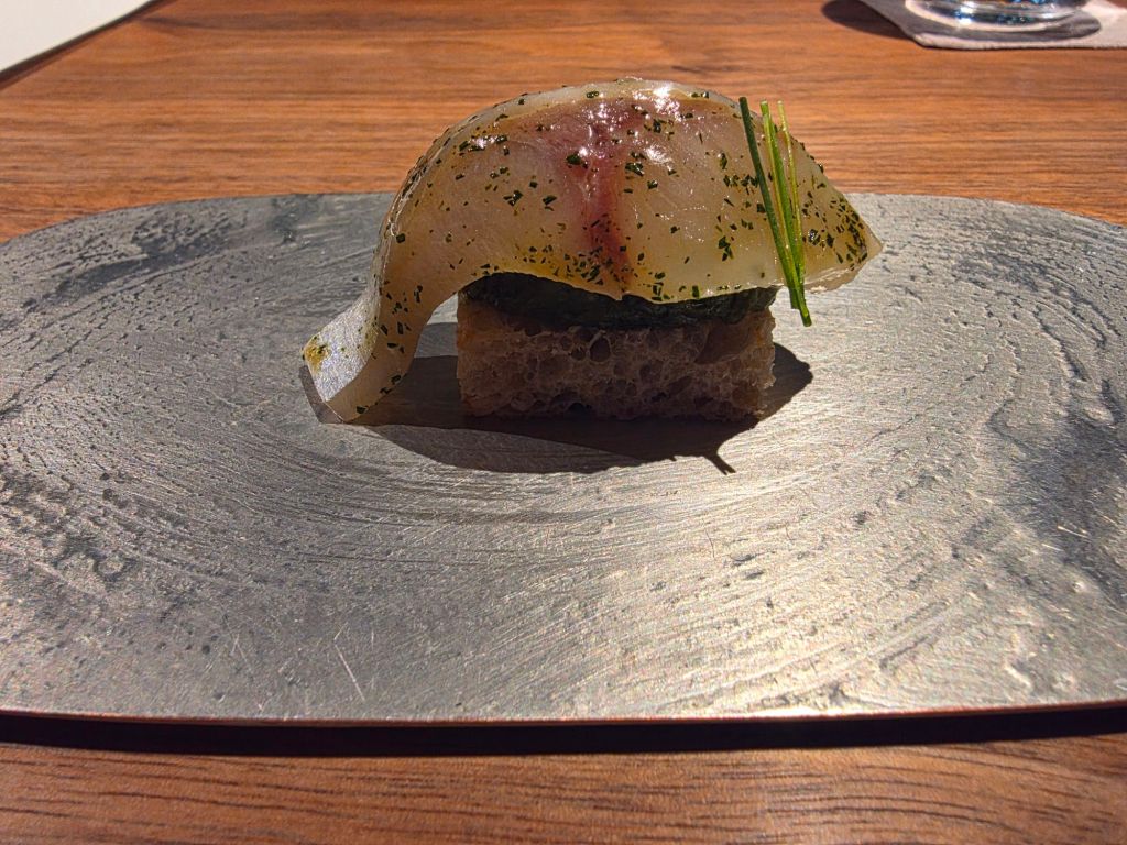 A piece of sushi featuring a translucent fish slice garnished with chives, placed on a small piece of textured bread on a metallic plate.