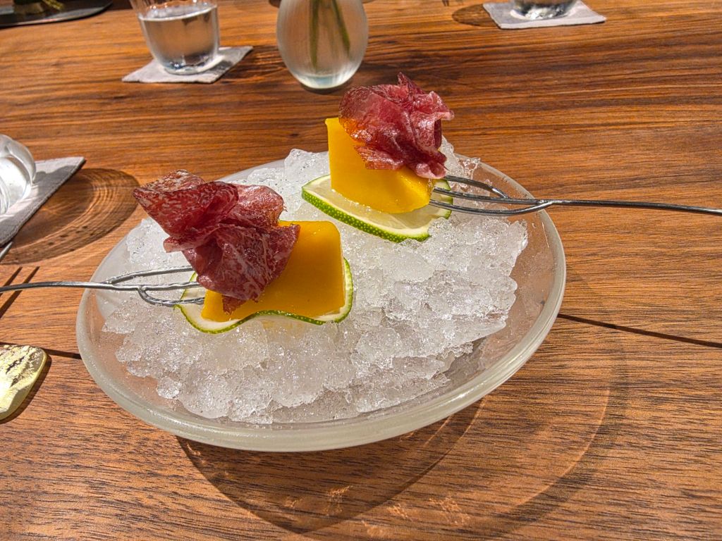 Two skewers with slices of cured meat and mango on a bed of crushed ice, served on a round plate.