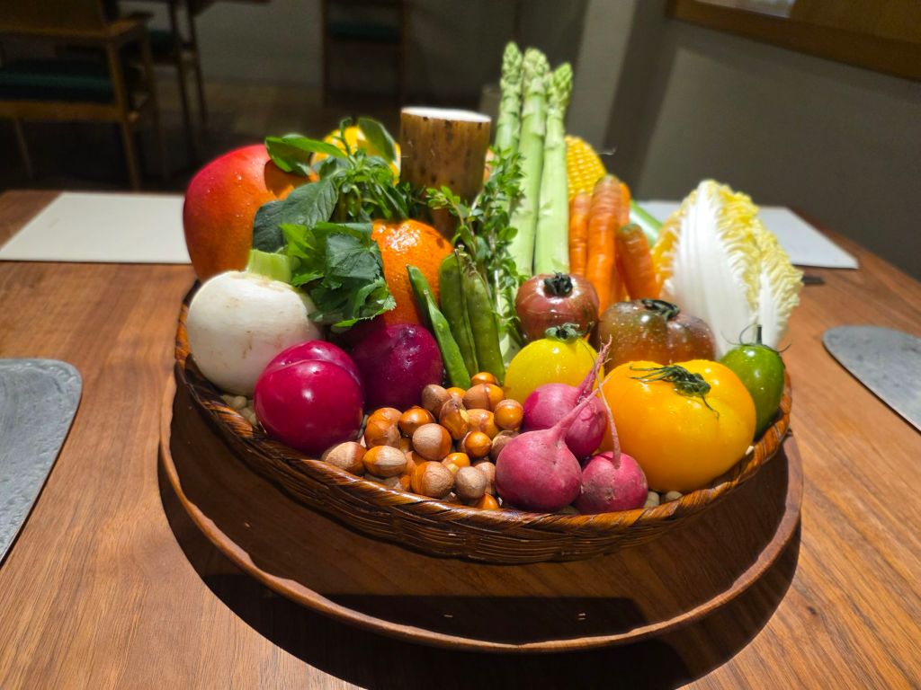 A vibrant assortment of fresh vegetables and fruits arranged in a woven basket, including apples, radishes, carrots, asparagus, various tomatoes, and greens.