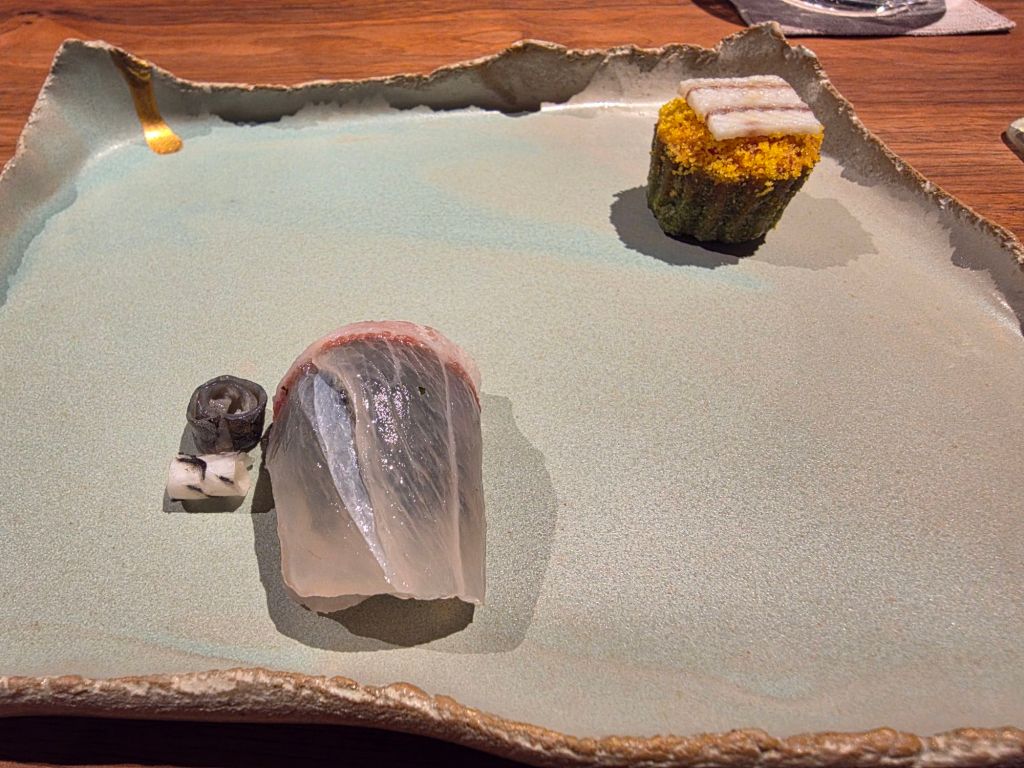 A plate featuring an artistic presentation of sushi, including a translucent slice of fish, a small piece of dark sushi, and a yellow-accented sushi roll.