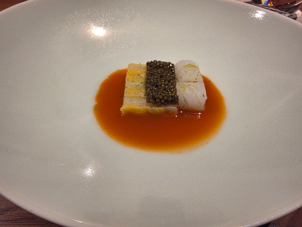 A gourmet dish featuring a piece of white fish topped with caviar, served in a light orange broth, garnished with herbs on a white plate.