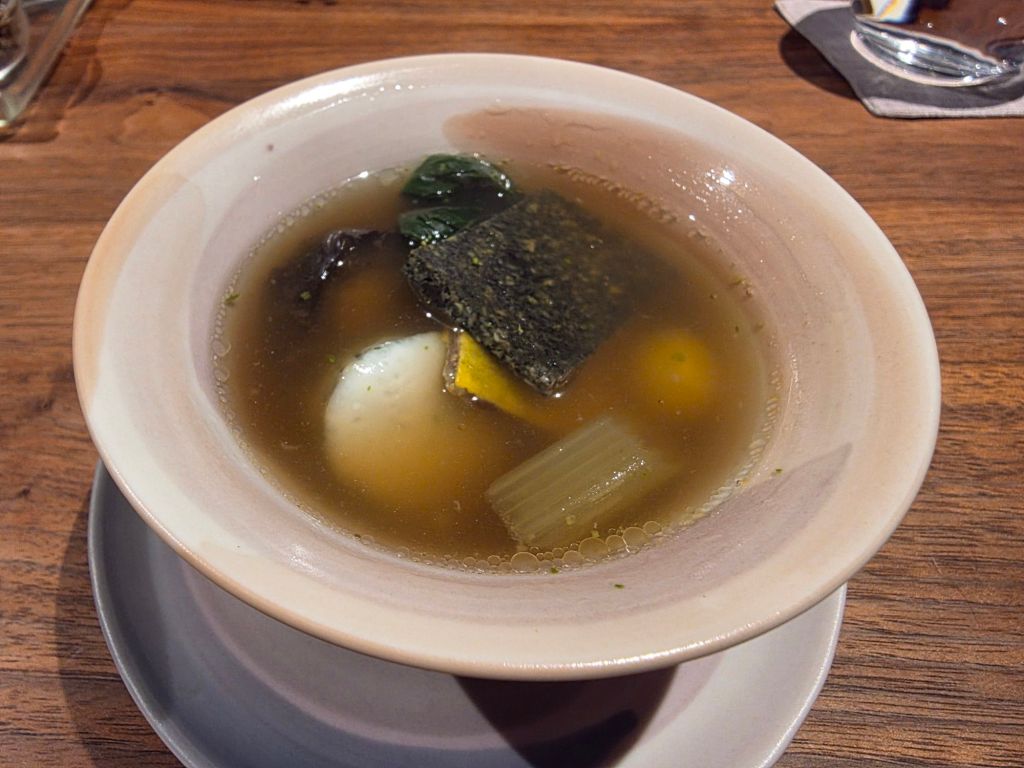 A bowl of clear broth soup containing a soft-boiled egg, seaweed, vegetables, and a piece of tofu on a wooden table.