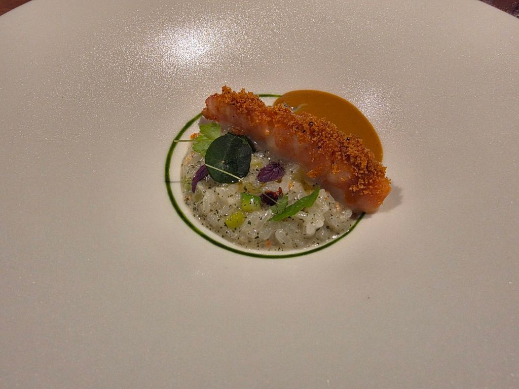 A gourmet dish featuring a crispy shrimp atop a bed of rice garnished with microgreens and a drizzle of sauce on a white plate.