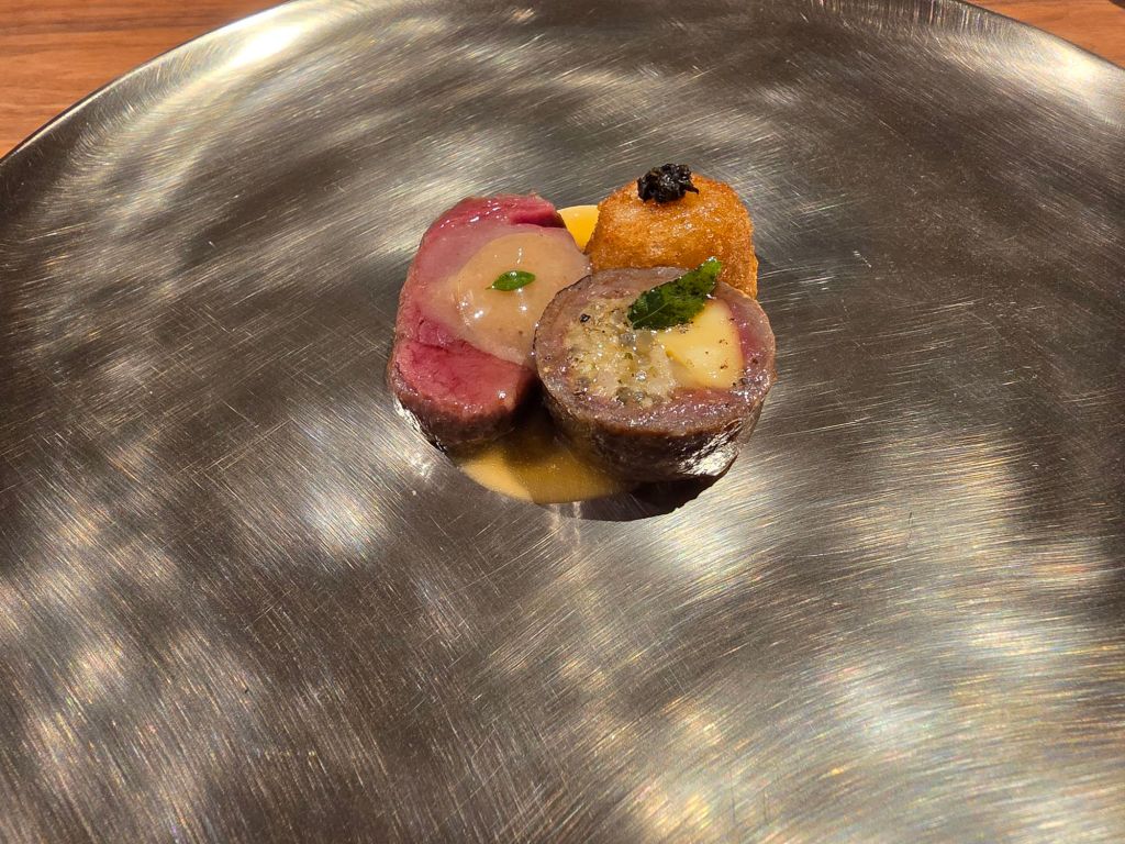 A gourmet plate featuring three pieces of meat: a cut of pink beef with a creamy sauce, a rolled meat preparation with a slice of butter, and a golden fried ball, garnished with a green leaf.