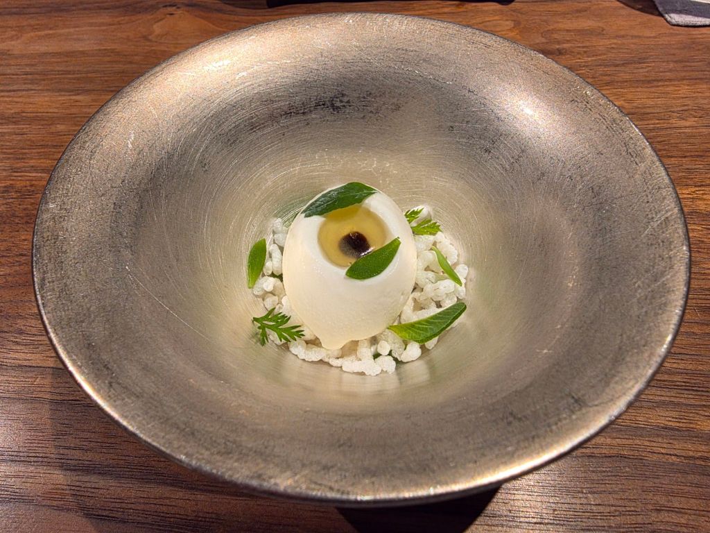 A minimalist gourmet dish featuring a white spherical centerpiece on a bed of small white crystals and garnished with fresh green leaves and a dark sauce.