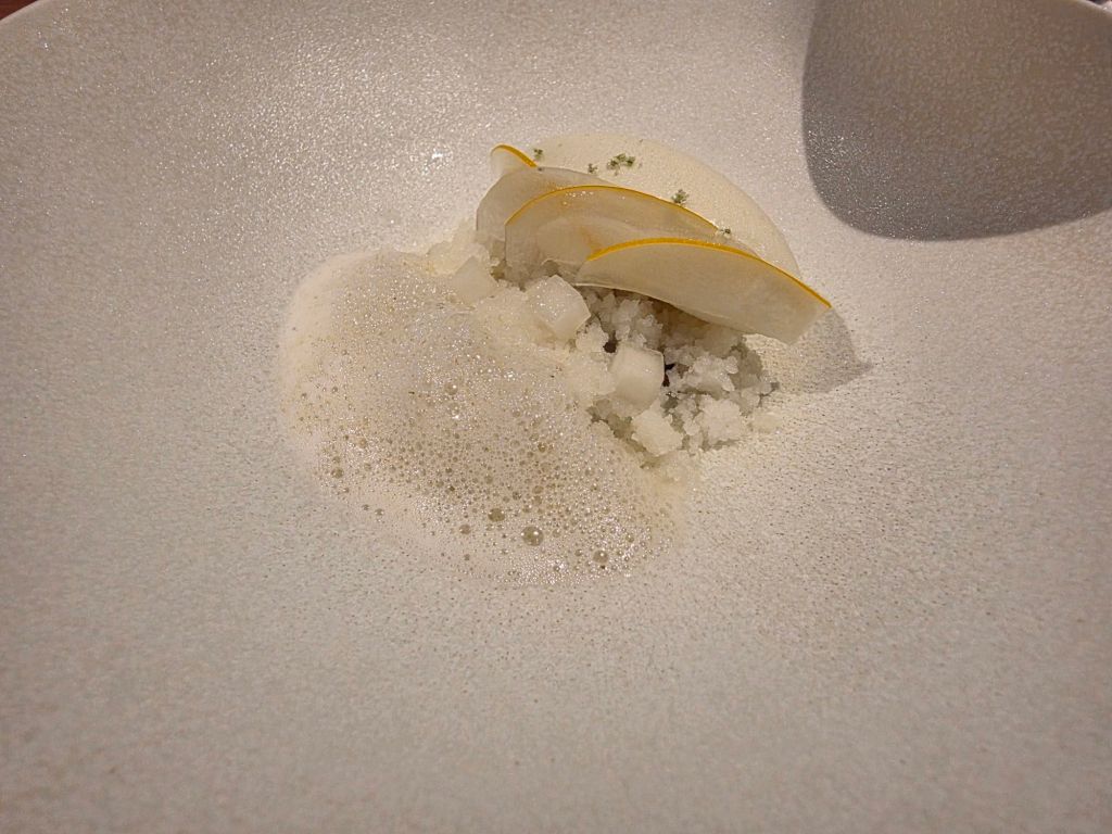 A white dish garnished with thin apple slices and a foam, featuring a bed of crushed ice or granita.
