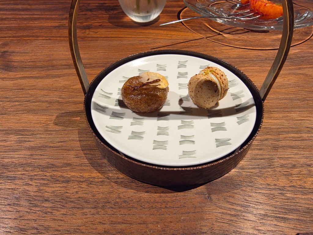 A round plate with a decorative pattern, featuring two gourmet bites: one resembling a small cake topped with an almond slice and the other a macaroon with a textured surface.