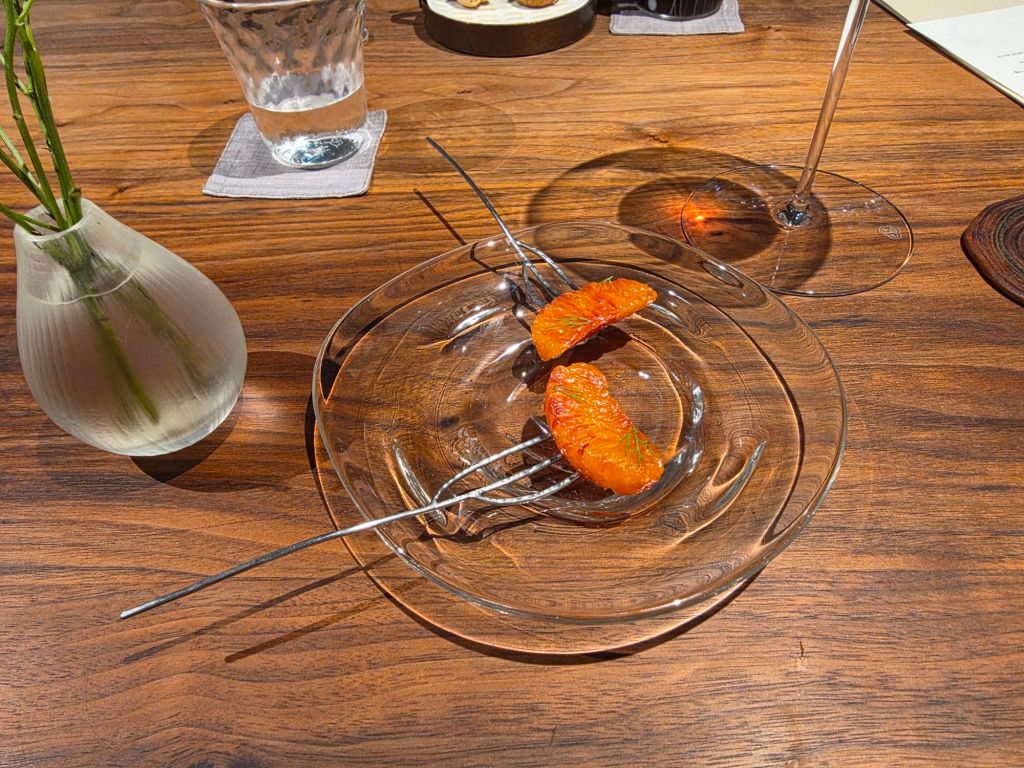 A clear glass plate with two orange fruit segments garnished with green, placed on a wooden table next to a small vase with a green plant and a glass of water.