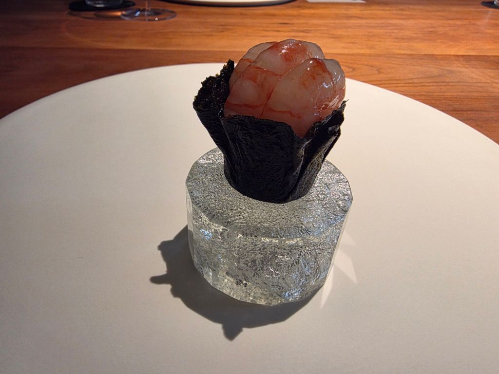 A gourmet dish featuring a piece of seafood topped with translucent shrimp on a bed of dark seaweed, presented on a clear, textured glass base.