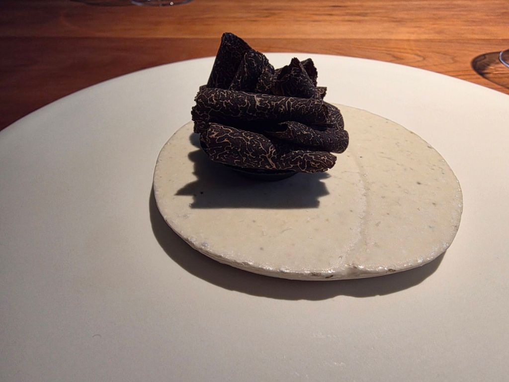 A gourmet dish featuring a mound of dark truffles placed on a smooth, circular white plate, with a wooden table surface in the background.
