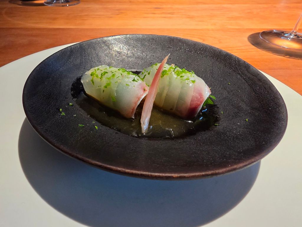 A close-up of a dark plate featuring two pieces of sushi, garnished with green herbs and served with a drizzle of sauce.