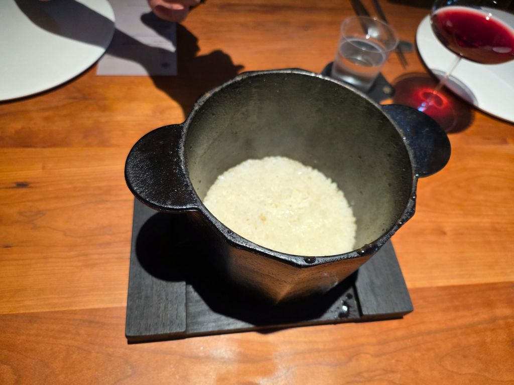 A steaming pot of rice served on a wooden table, with a glass of red wine and water in the background.