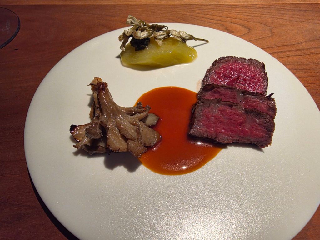 A gourmet dish featuring slices of roasted meat, accompanied by a vibrant sauce, a piece of pickled vegetable, and a cluster of sautéed mushrooms, all presented on a white plate.