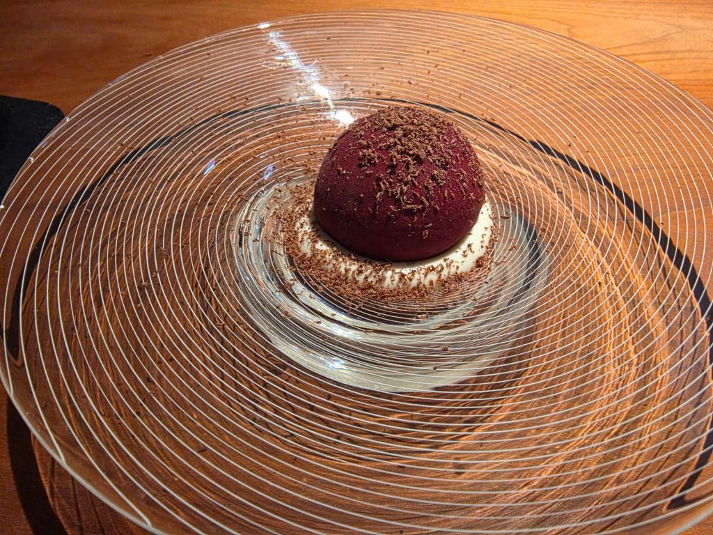 A round chocolate dessert topped with shavings, sitting on a circular clear plate with concentric lines.