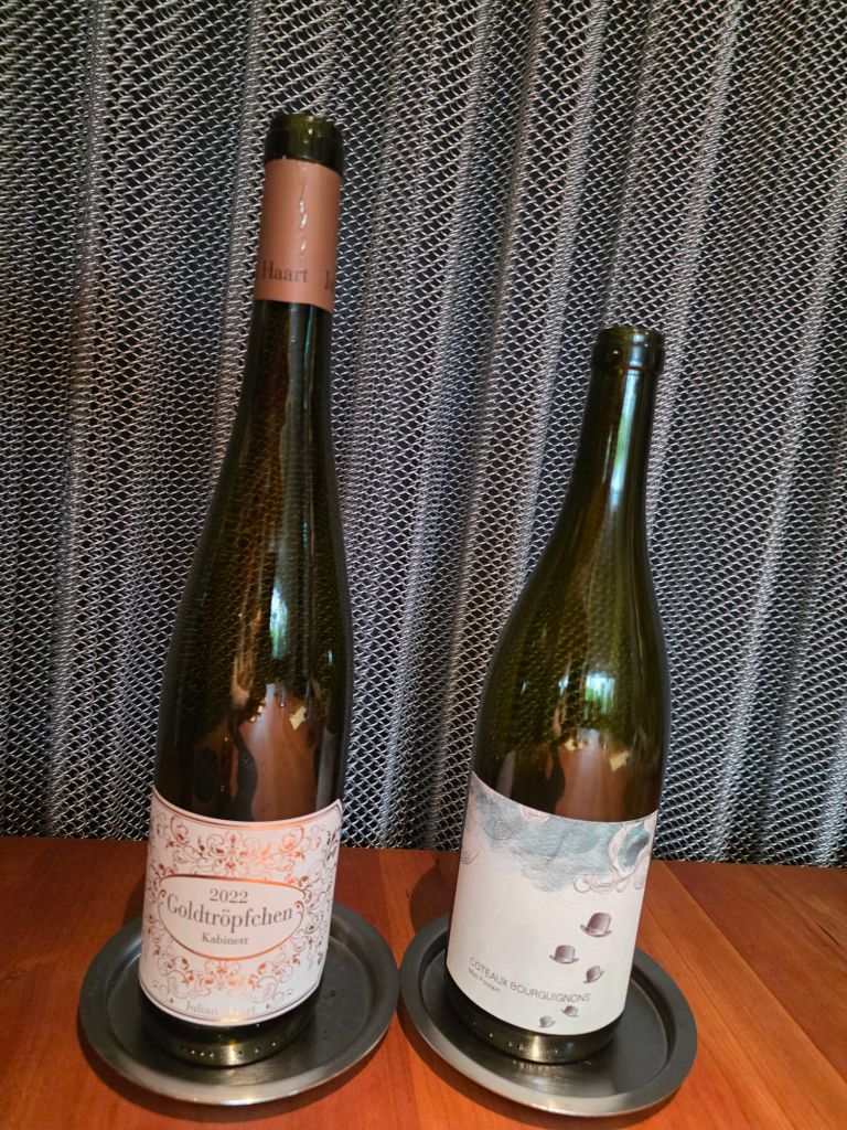 Two wine bottles placed on metal coasters. The left bottle features a label reading '2022 Goldtröpfchen Kabinett,' while the right bottle has a label that says 'Coteaux Bourguignons.' The background consists of a textured mesh material.