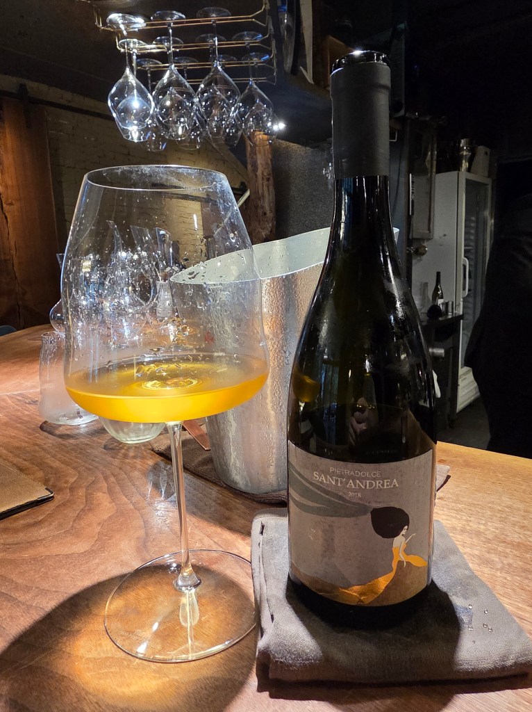 A glass of amber-colored wine sits next to a bottle labeled 'Pietradolce Sant'Andrea 2018' on a wooden table, with a backdrop of hanging wine glasses.